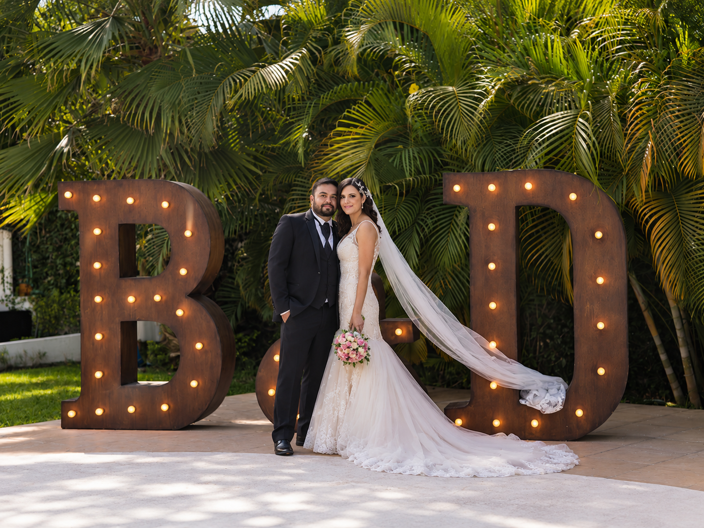 Novios junto a letras gigantes iluminadas con sus iniciales entre palmeras en Jardín Allegra 55, Cuernavaca