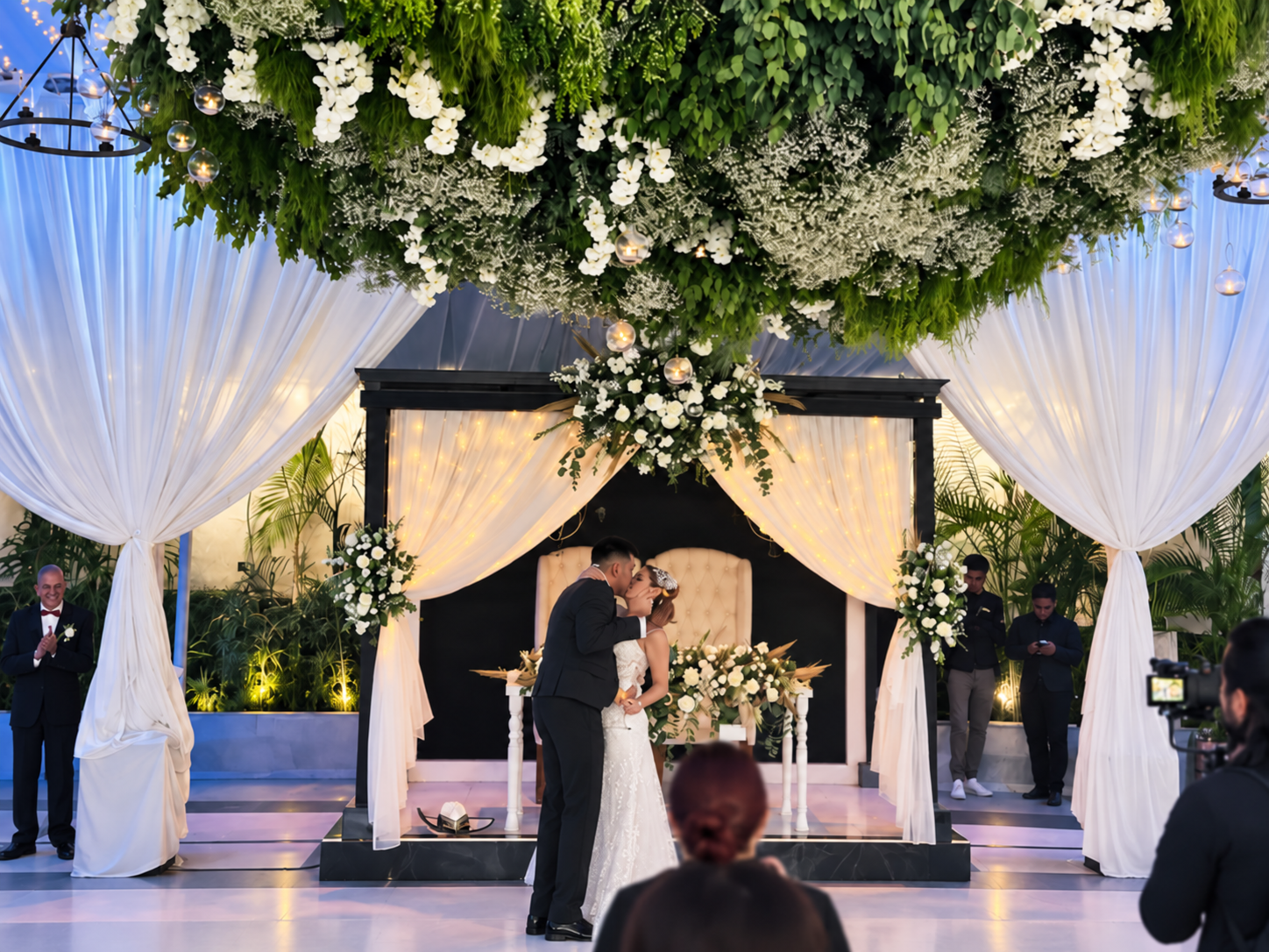 Beso de novios bajo pérgola con techo floral blanco durante su ceremonia en Jardín Allegra 55