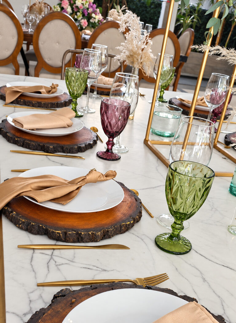 Montaje de mesa con bases de madera, copas verdes y moradas y cubiertos dorados en Jardín Allegra 55, Cuernavaca