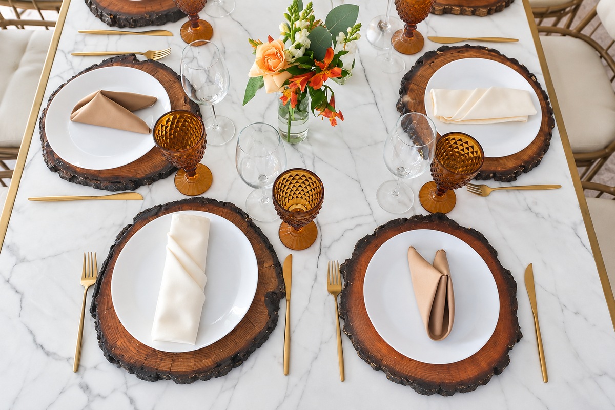 Montaje de mesa con bases de madera, platos blancos, copas ámbar y cubiertos dorados en Jardín Allegra 55