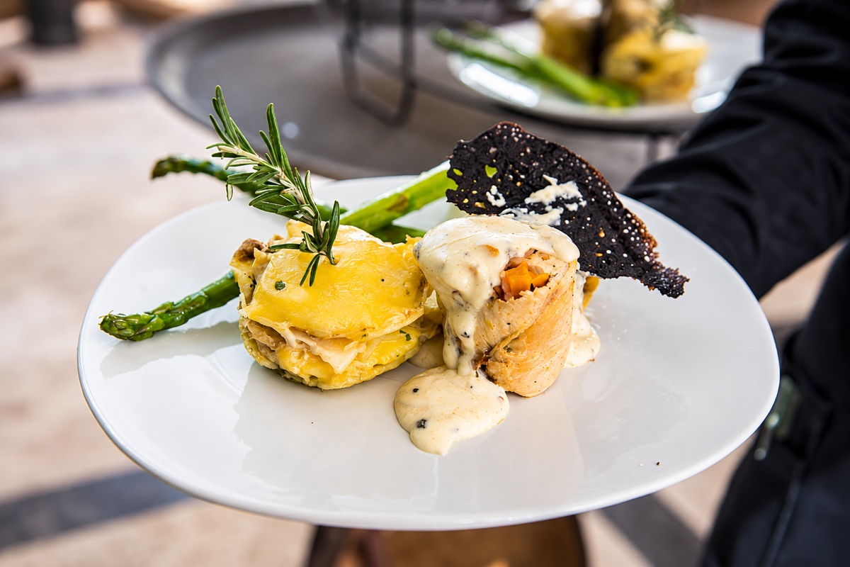 Pollo relleno con salsa cremosa, guarnición dorada con romero, espárragos y una lámina crujiente oscura sobre plato blanco.